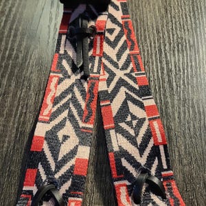 Op de afbeelding: Een patroonband met een zwarte gesp en metalen ringen. De band heeft een herhalend geometrisch ontwerp in rood, zwart en gebroken wit. De band is gemaakt van een geweven materiaal.