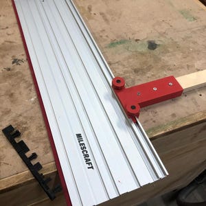 Puede incluir: Una guía de corte Milescraft plateada y roja con una guía ajustable roja y un accesorio de plástico negro. La guía está diseñada para cortes precisos en proyectos de carpintería. El logotipo de Milescraft está impreso en el lateral.