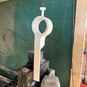 May include: A white, wooden, custom-made object with a circular opening and a screw at the top. The object is mounted on a black machine with a gray glove.