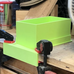 May include: A bright green rectangular box is clamped to a wooden surface. The box is made of a solid material and has a simple, modern design. Two black clamps with red accents secure the box in place.