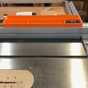 May include: An orange plastic organizer with multiple compartments sits on a silver table saw. A black remote control is in one of the compartments. The saw blade is visible, along with the saw's controls.