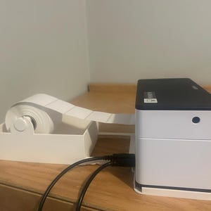 Op de afbeelding: Een wit en zwarte labelprinter met een rol blanco labels in een witte houder. De printer is aangesloten op een USB-kabel. De printer staat op een houten oppervlak.
