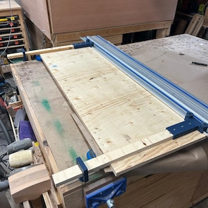 May include: A woodworking scene featuring a light-colored plywood board clamped to a workbench. A blue and silver guide rail is positioned along the edge of the board, with additional clamps securing the wood. The workbench is made of wood.