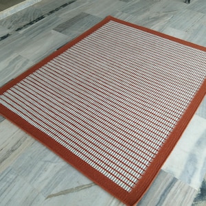 May include: A rectangular area rug featuring a grid pattern of white horizontal lines set against a deep terracotta orange background. The rug has a solid terracotta orange border that frames the central patterned section, creating a clean edge.