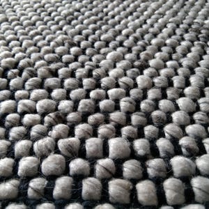 May include: Close-up of a grey and black textured rug. The rug features a repeating pattern of rounded, woven elements, creating a tactile surface. The colour palette is primarily neutral, with variations in grey and black.