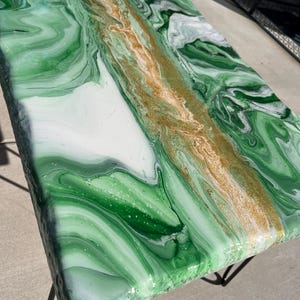 May include: A rectangular table with a green, white, and gold marbled epoxy resin surface. The surface has a glossy finish and is supported by black metal legs. The design features swirling patterns of green and white with a gold stripe down the center.