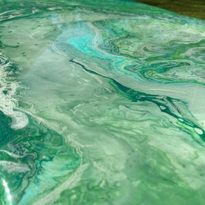 May include: Close-up of a surface with a fluid, abstract design. The color palette features shades of green and white, creating a marbled effect. The glossy finish reflects light, enhancing the depth and texture of the artwork.
