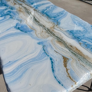 May include: Rectangular table with a resin top featuring a swirling design in shades of white, light blue, and gold. The table has a unique, marbled appearance. The legs are black.