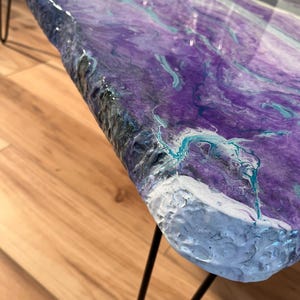 May include: A live edge coffee table with a vibrant, swirled epoxy resin top in shades of purple, teal, and white. The table has black hairpin legs and a glossy finish. The table's dimensions are approximately 48 inches long, 24 inches wide, and 18 inches high.