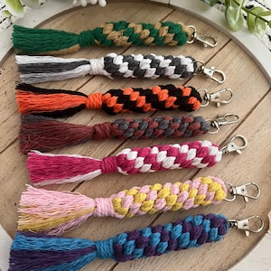Puede incluir: Una colección de llaveros de macramé hechos a mano con diseños trenzados y borlas coloridas. Cada llavero presenta una combinación de colores diferente, incluyendo verde, blanco y negro, naranja y negro, y rosa.