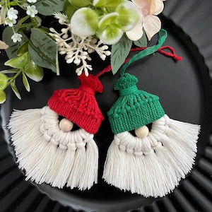 May include: Two Christmas ornaments shaped like gnomes. One gnome has a red knitted hat and the other has a green knitted hat. Both have white macrame beards and wooden bead noses. The ornaments are on a black plate with greenery.