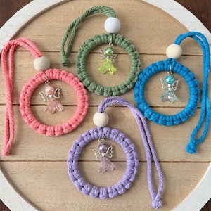 May include: Five macrame angel ornaments in pink, green, blue, and purple. Each circular ornament has a small angel charm and a wooden bead at the top. The ornaments are displayed on a wooden tray.