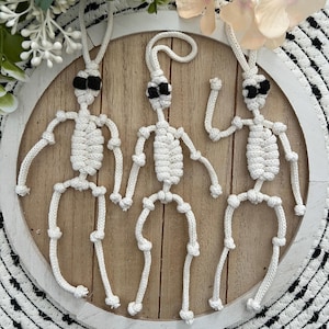 May include: Three handmade macrame skeleton figures with black eye coverings are displayed on a wooden round board. The skeletons are made of white rope and have articulated arms and legs. The board is set on a black and white patterned surface, with floral accents.