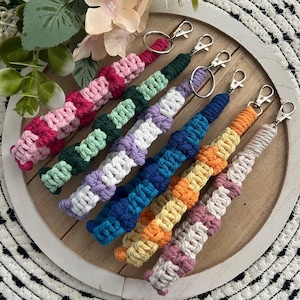 May include: A collection of colorful macrame keychains in various shades, including pink, green, purple, blue, orange, and beige. Each keychain features a woven design and a silver-tone clasp. The keychains are arranged on a wooden tray.