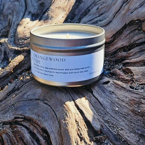 May include: A silver metal candle tin with a white label that reads "ORANGEWOOD TIDE". The candle is on a piece of driftwood. The label describes the candle's scent as a blend of citrus, sea-washed wood, and sun-bleached birch.