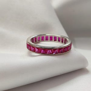 Könnte beinhalten: Ein silberner Ring mit einem durchgehenden Band aus quadratisch geschliffenen, leuchtend rosa Edelsteinen. Der Ring ist vor einem weißen Hintergrund platziert, der die Farbe der Edelsteine und das Design des Rings hervorhebt. Ein Schmuckstück.