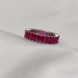 Op de afbeelding: Een zilverkleurige ring met een doorlopende band van rechthoekige, levendige roze edelstenen. De ring is geplaatst tegen een witte, getextureerde achtergrond, die de glans van de edelstenen benadrukt. Dit is een modeaccessoire.
