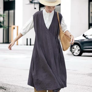 Pode incluir: Uma mulher vestindo um vestido envelope cinza escuro sobre uma blusa branca, combinada com shorts bege e sandálias pretas. Ela também está usando um chapéu de palha e carregando uma bolsa de tecido. A roupa é adequada para o clima quente.