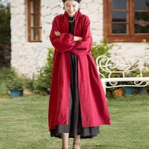 Può includere: Una donna indossa un lungo cappotto rosso fluente sopra un vestito scuro, in piedi sull'erba verde. Il cappotto ha maniche lunghe e una vestibilità ampia. Lo sfondo include una casa in pietra e una panchina bianca.