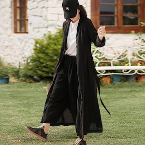 以下が含まれることがあります： 黒いリネンのダスターコート、黒い野球帽、パンツ。白いTシャツと茶色のソールが付いた黒いスニーカーが含まれています。石造りの建物の前で、緑の芝生を歩いている人。