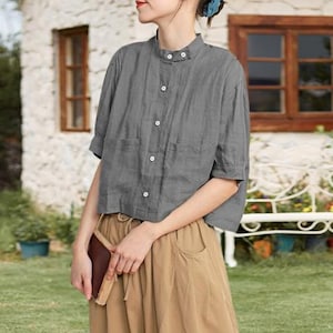 Könnte beinhalten: Ein graues kurzärmeliges Hemd mit Stehkragen und Knopfleiste. Das Hemd hat zwei Vordertaschen und wird mit einer beigefarbenen Hose kombiniert. Das Outfit ist lässig und für warmes Wetter geeignet.