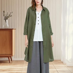 May include: An olive green long-sleeve shirt worn over a white top with a V-neck and button closure. The outfit includes gray culottes and brown sandals. The background features a wooden cabinet, a vase with plants, and a light-colored wall.