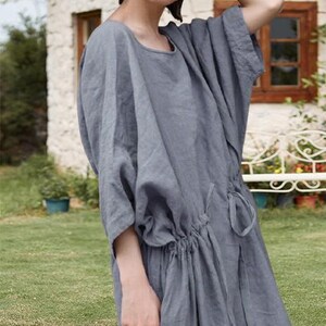 May include: A gray linen dress with a round neckline and short, wide sleeves. The dress features a drawstring waist and falls to the mid-calf. The model is standing outside on a sunny day.