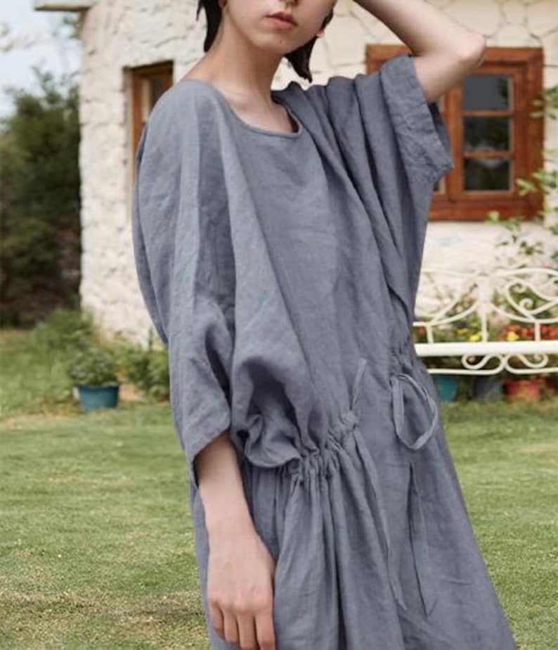 May include: A gray linen dress with a round neckline and short, wide sleeves. The dress features a drawstring waist and falls to the mid-calf. The model is standing outside on a sunny day.