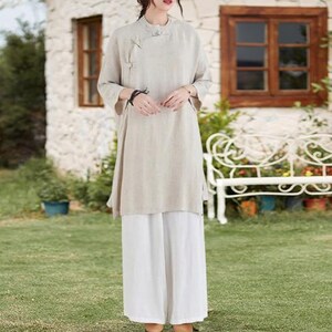 May include: A beige linen tunic with a mandarin collar and side slits, paired with wide-leg white pants. The tunic features a button closure on the side. The outfit is suitable for casual wear.