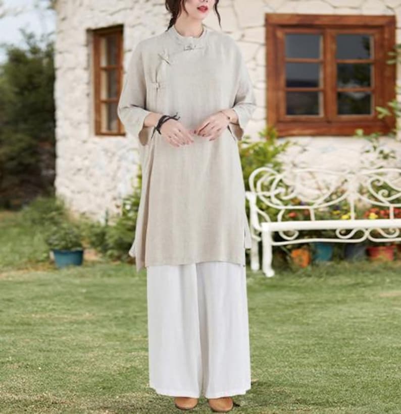 May include: A beige linen tunic with a mandarin collar and side slits, paired with wide-leg white pants. The tunic features a button closure on the side. The outfit is suitable for casual wear.