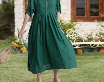 May include: A green linen dress with a V-neckline and button details. The dress has short sleeves and a gathered waist, and is paired with yellow shoes. The person is holding a bouquet of yellow flowers and a woven bag.