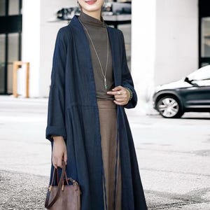 May include: A long, navy blue linen duster coat with a relaxed fit. The coat is open in the front and has long sleeves. The model is wearing a brown turtleneck, a long brown skirt, and a brown leather handbag. A silver necklace with a pendant is visible.
