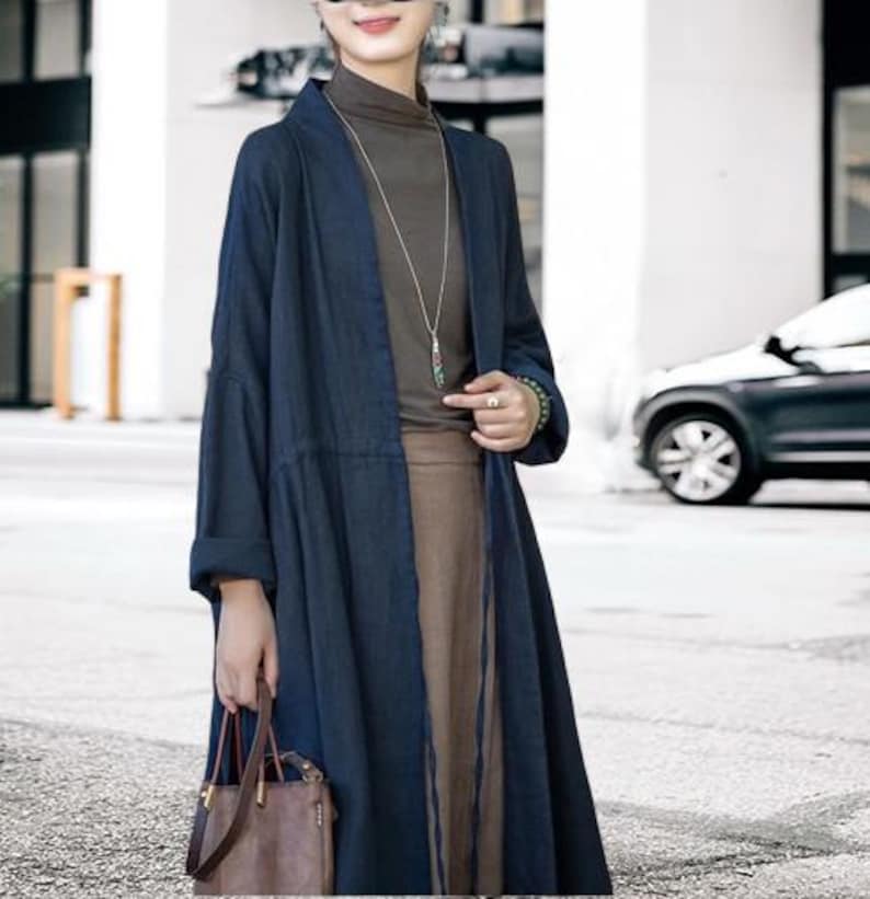 May include: A long, navy blue linen duster coat with a relaxed fit. The coat is open in the front and has long sleeves. The model is wearing a brown turtleneck, a long brown skirt, and a brown leather handbag. A silver necklace with a pendant is visible.