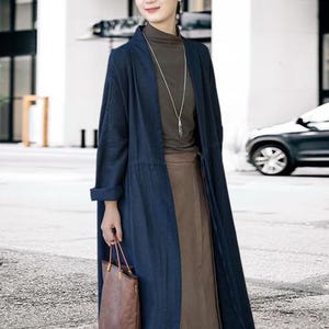 May include: A long, navy blue linen coat with a matching belt, worn over a brown top and skirt. The coat has long sleeves and a relaxed fit. A brown leather tote bag is held in the hand. A silver necklace is worn.