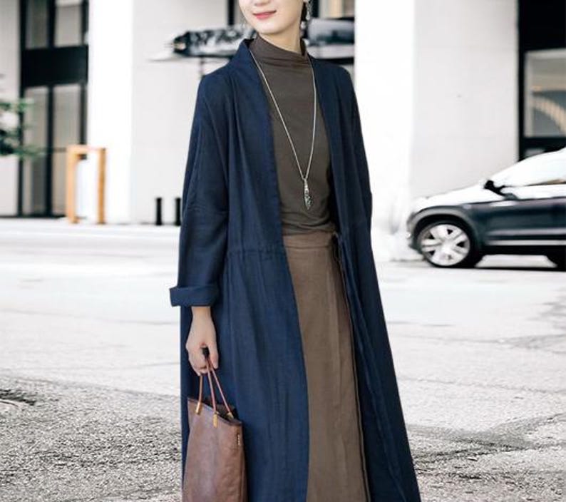 May include: A long, navy blue linen coat with a matching belt, worn over a brown top and skirt. The coat has long sleeves and a relaxed fit. A brown leather tote bag is held in the hand. A silver necklace is worn.