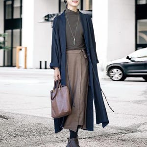May include: A navy blue long coat with a matching tie, worn over a brown skirt and a dark green turtleneck. A brown leather tote bag is held in the hand. The outfit is completed with black leggings and brown boots.