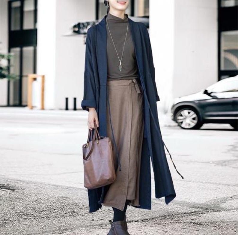 May include: A navy blue long coat with a matching tie, worn over a brown skirt and a dark green turtleneck. A brown leather tote bag is held in the hand. The outfit is completed with black leggings and brown boots.