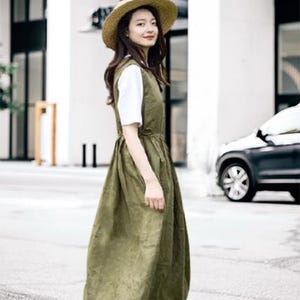 May include: An olive green dress with a white undershirt and a straw hat. The dress has a sleeveless design and a gathered waist. The model is wearing brown sandals. The background includes a building and a car.