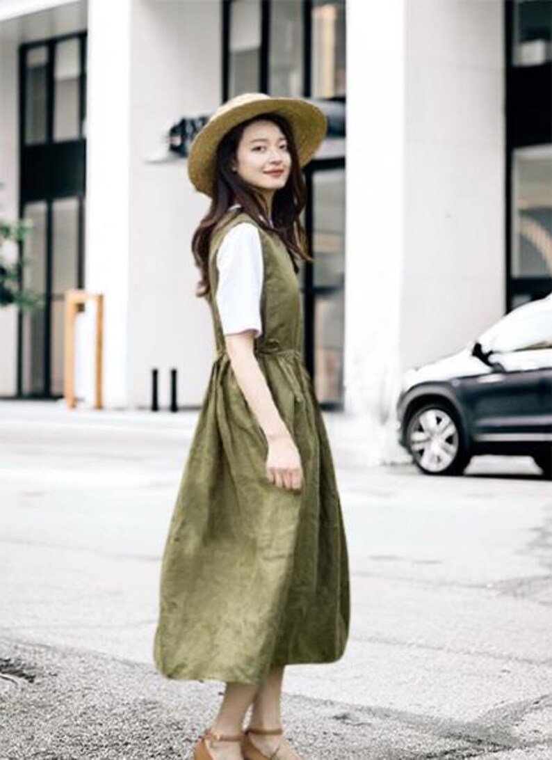 May include: An olive green dress with a white undershirt and a straw hat. The dress has a sleeveless design and a gathered waist. The model is wearing brown sandals. The background includes a building and a car.