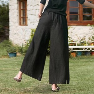 May include: Black wide-leg trousers with an elasticated waistband. The trousers are made of a lightweight fabric and have a relaxed fit. The trousers are shown outdoors on a grassy lawn.