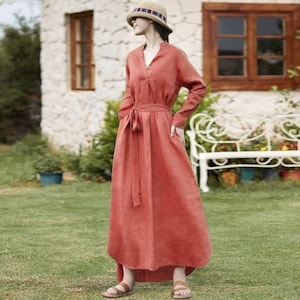 May include: A long, rust-colored dress with long sleeves and a V-neck. The dress features a tie belt at the waist and pockets. The person is wearing a straw hat with a striped band and brown sandals. The background includes a stone building and green grass.