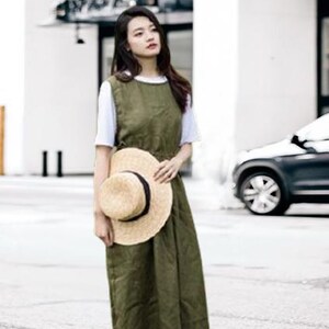 May include: An olive green sleeveless dress worn over a white t-shirt. A straw hat with a black band is held in front of the dress. The outfit is worn outdoors on a sunny day.