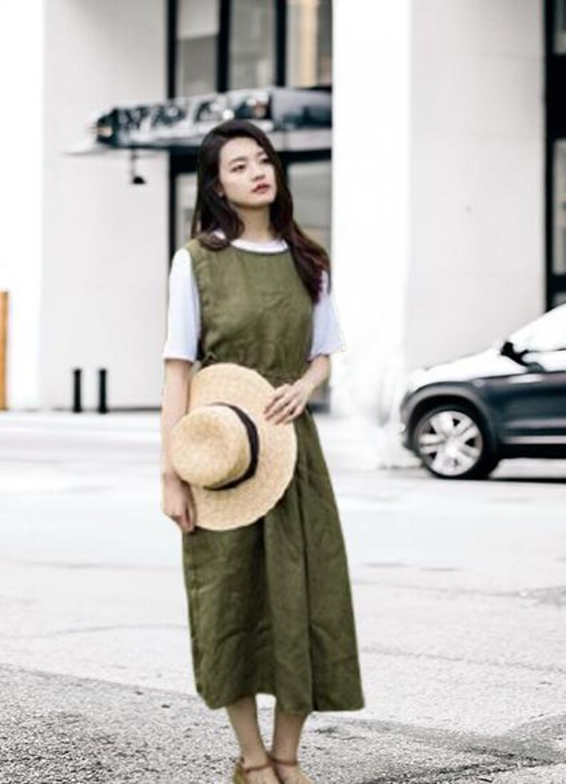 May include: An olive green sleeveless dress worn over a white t-shirt. A straw hat with a black band is held in front of the dress. The outfit is worn outdoors on a sunny day.