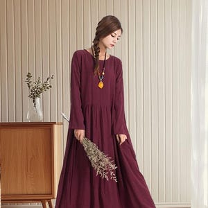 May include: A full-length, burgundy dress with long sleeves and a round neckline. The dress features a gathered waist and is worn with tan shoes. The person is holding dried flowers. A necklace with a yellow pendant completes the look. The dress is approximately 150 cm in length.