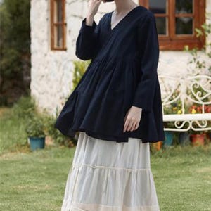 Könnte beinhalten: Ein Damenoutfit mit einem dunkelblauen V-Ausschnitt-Oberteil mit langen Ärmeln und einem cremefarbenen, gestuften Rock. Das Outfit wird durch einen Strohhut und schwarze Mary-Jane-Schuhe ergänzt. Der Hintergrund zeigt ein weißes Gebäude und grünes Gras.