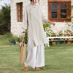 May include: A beige linen tunic with a mandarin collar and side slits, paired with wide-leg white pants. The outfit is accessorized with a woven tote bag and brown shoes. The ensemble is suitable for casual wear.