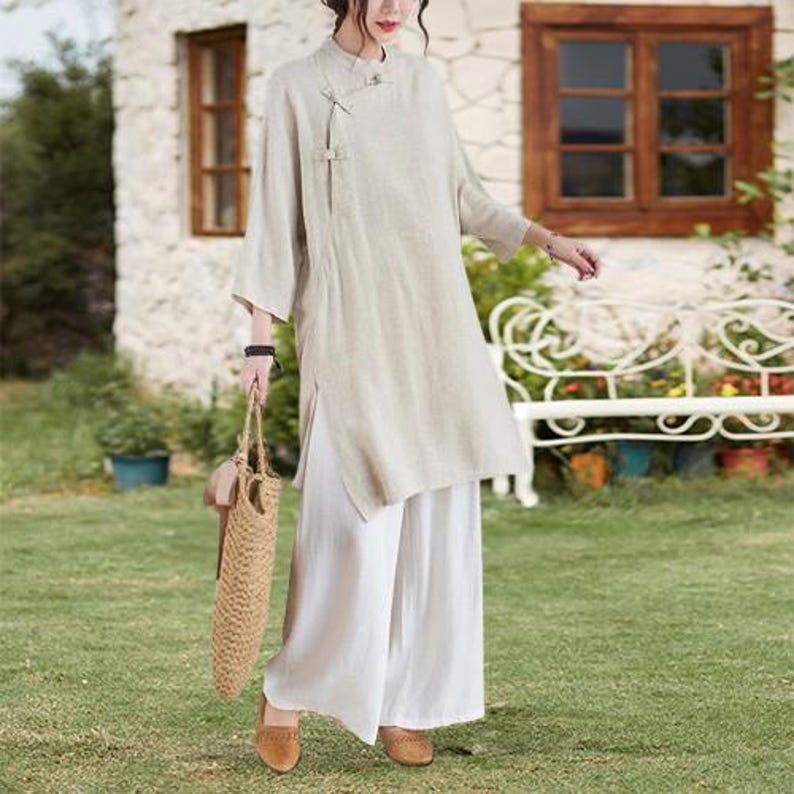 May include: A beige linen tunic with a mandarin collar and side slits, paired with wide-leg white pants. The outfit is accessorized with a woven tote bag and brown shoes. The ensemble is suitable for casual wear.