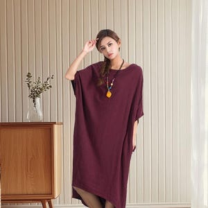 May include: A burgundy-colored, loose-fitting dress with short sleeves and a boat neckline. The dress falls to mid-calf length and has an asymmetrical hemline. A colorful necklace is worn with the dress. The dress is made of a soft, flowing fabric.