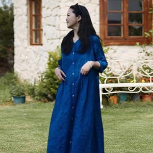 May include: A long, button-down, blue dress with a V-neck. The dress is worn outdoors in front of a stone building with wooden windows and a white bench. The dress has long sleeves and a flowing skirt.