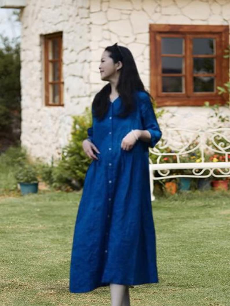May include: A long, button-down, blue dress with a V-neck. The dress is worn outdoors in front of a stone building with wooden windows and a white bench. The dress has long sleeves and a flowing skirt.